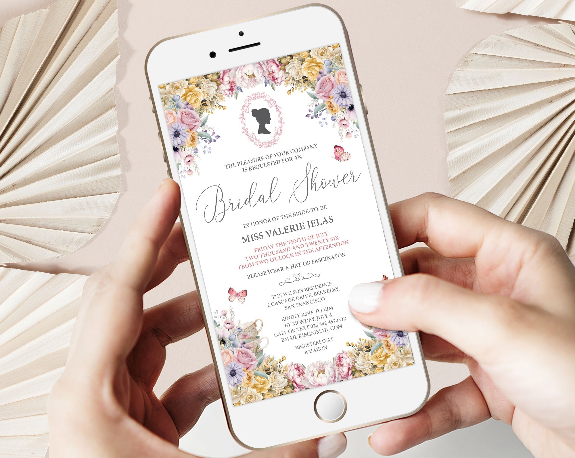 two hands holding a mobile phone with a pastel flower regency style bridal shower invitation showing on the screen, beige dried leaves in the background