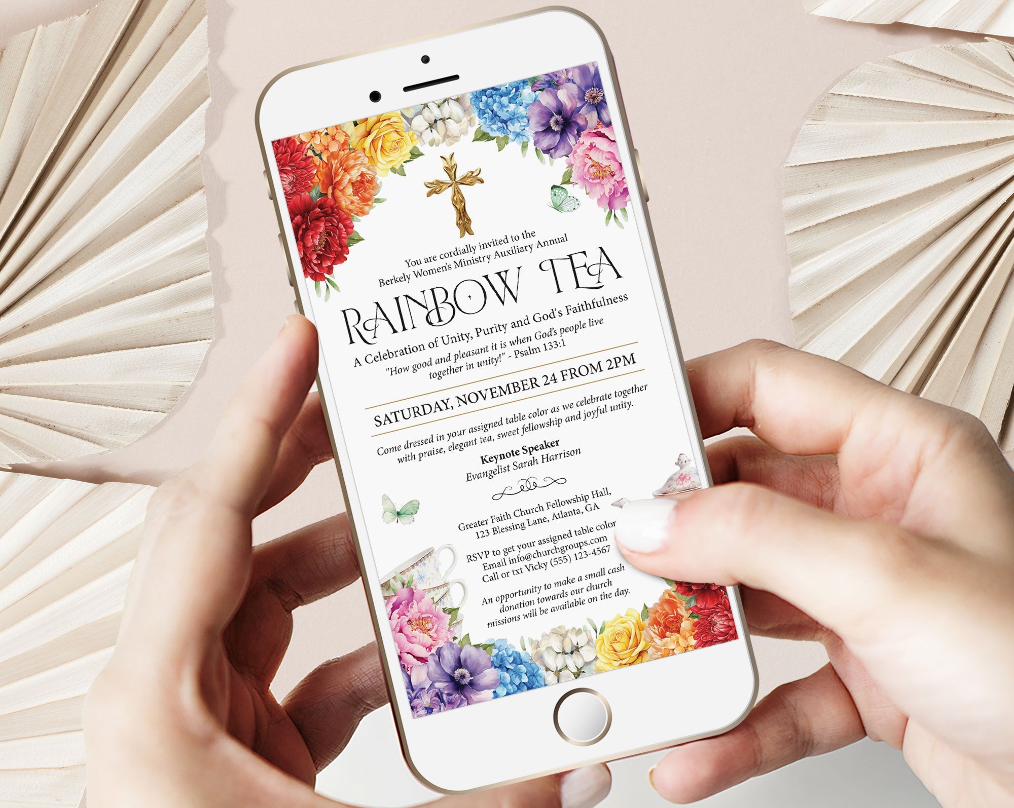 two hands holding a mobile phone showing a Church Rainbow Tea Invitation with bright Flowers, gold cross and Teapot with a beige background and two dried leaves