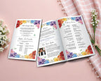 An open 11 x 8.5 folded Rainbow tea church program booklet displayed on a soft pink surface, surrounded by white flowers and a pink plaid cloth. The booklet has colorful floral watercolor borders and printed ceremony text.
