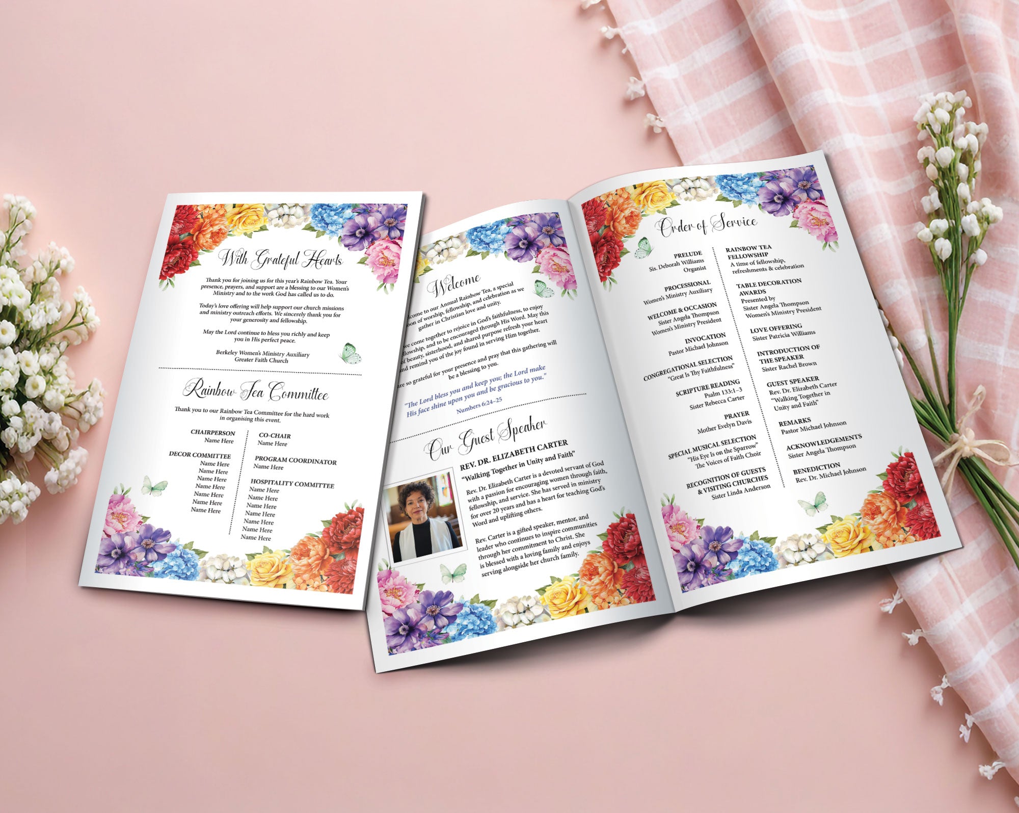 An open 11 x 8.5 folded Rainbow tea church program booklet displayed on a soft pink surface, surrounded by white flowers and a pink plaid cloth. The booklet has colorful floral watercolor borders and printed ceremony text.