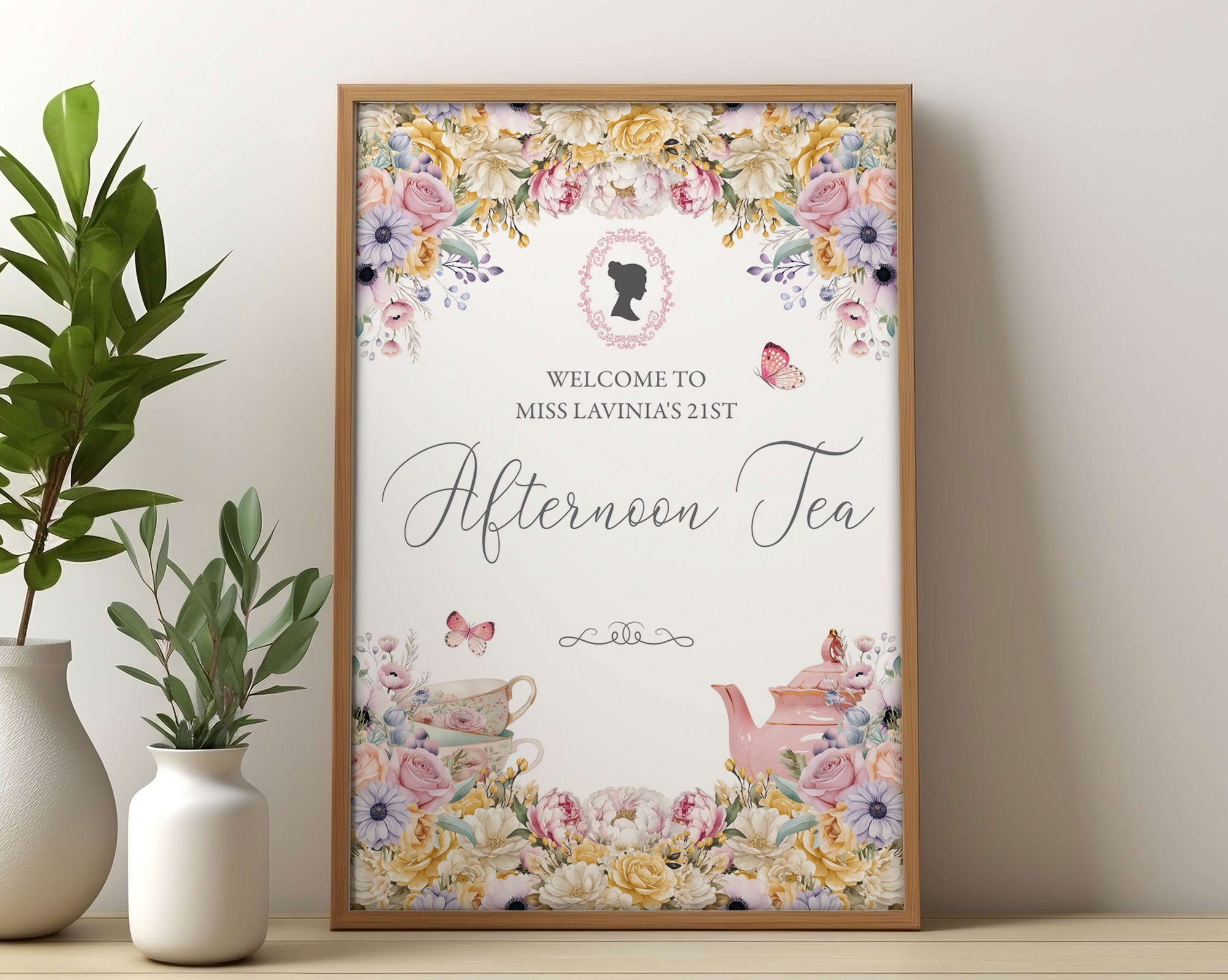 Small Bridgerton inspired afternoon tea digital welcome sign in a wooden frame leaning up against a beige wall next to some greenery in vases