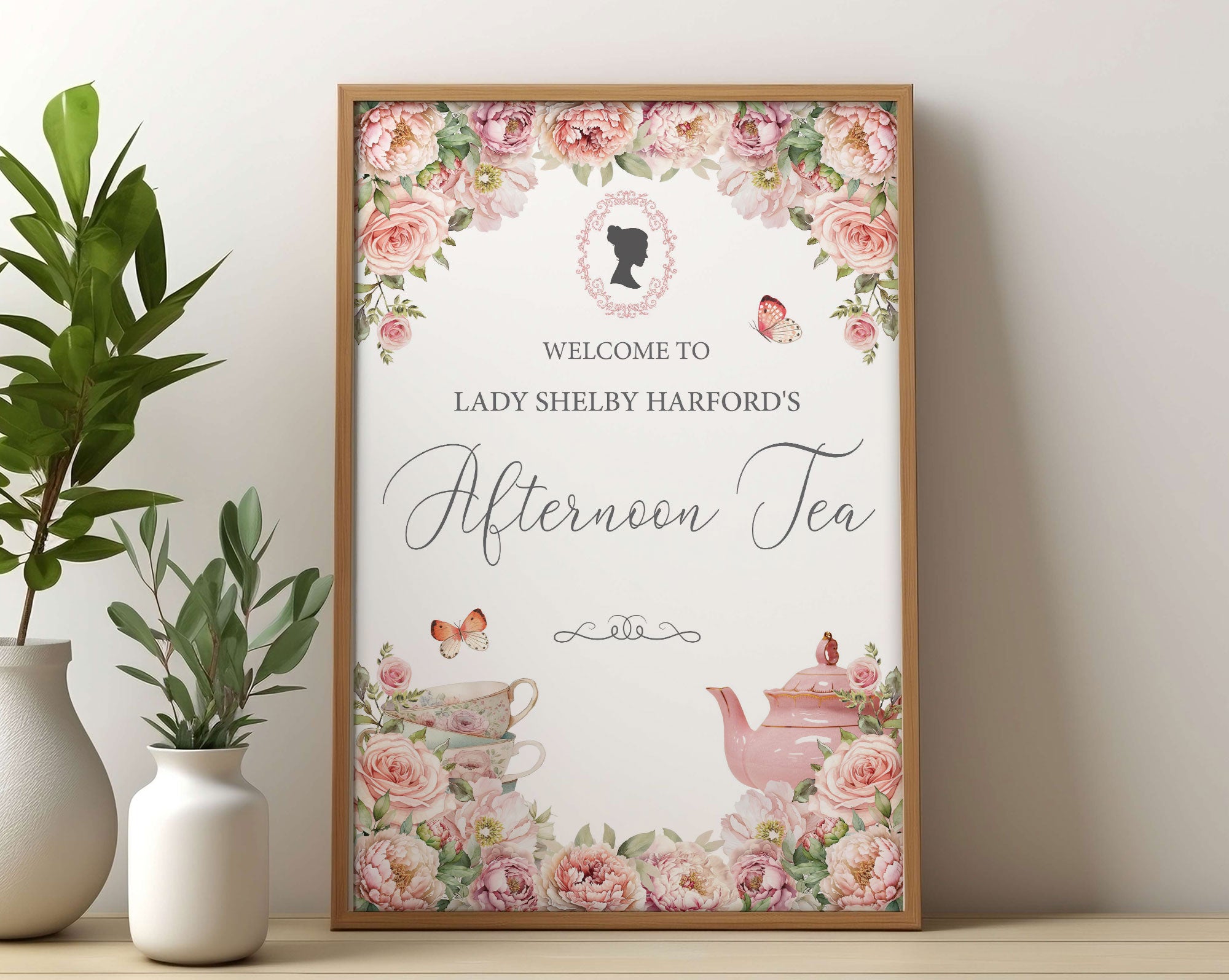 pretty pink flower sign for an Afternoon Tea with regency style cameo, teapot and butterflies in a wooden frame leaning against a beige wall next to greenery in a vase