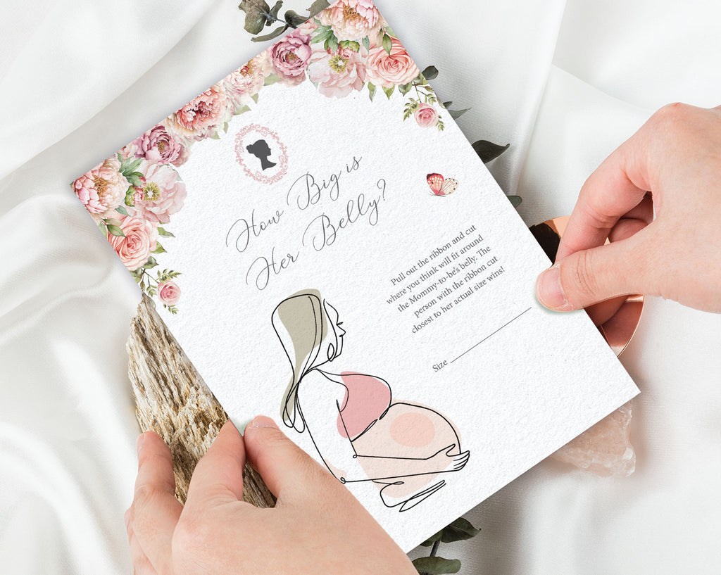 two hands holding a 'how big is her belly' baby shower game card with a white silk cloth in the background