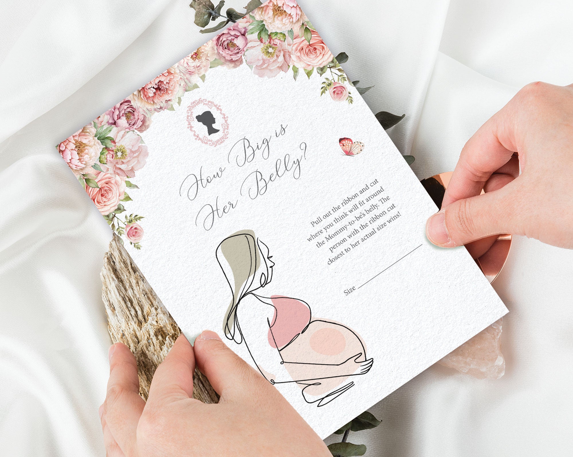 two hands holding a 'how big is her belly' baby shower game card with a white silk cloth in the background