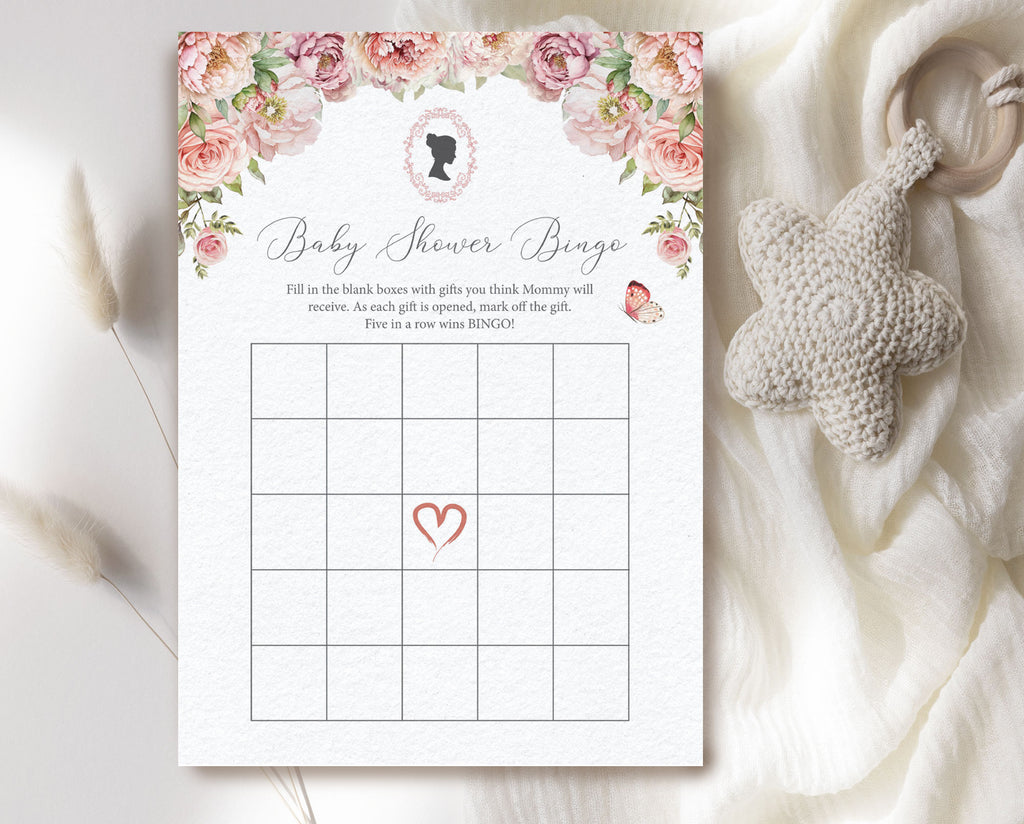 a blush pink regency style 'baby shower bingo' card laying on a cream cloth with a baby teether next to it