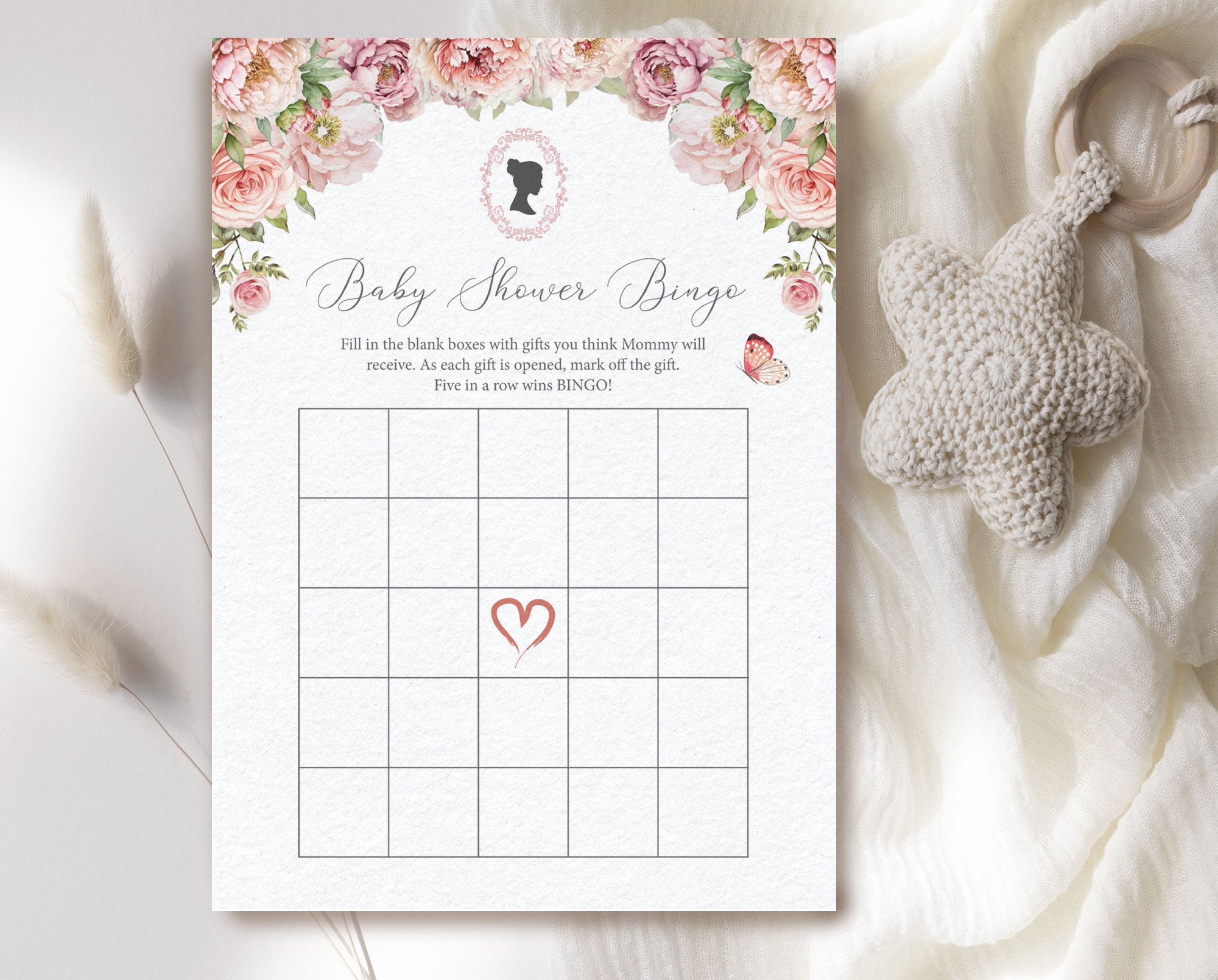 a blush pink regency style 'baby shower bingo' card laying on a cream cloth with a baby teether next to it