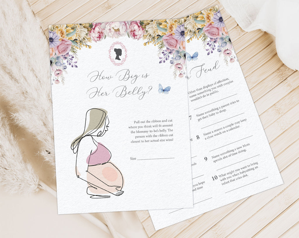 2 Pastel baby shower game cards laying on a wooden background next to a cloth and some dried plants