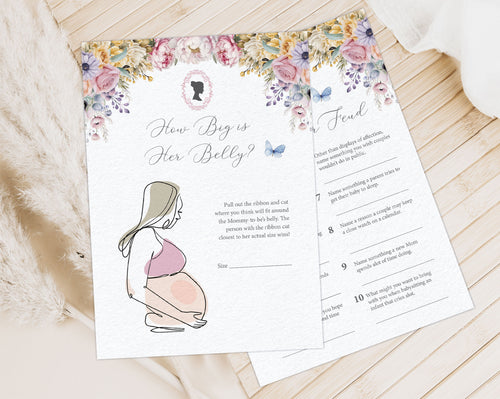 2 Pastel baby shower game cards laying on a wooden background next to a cloth and some dried plants