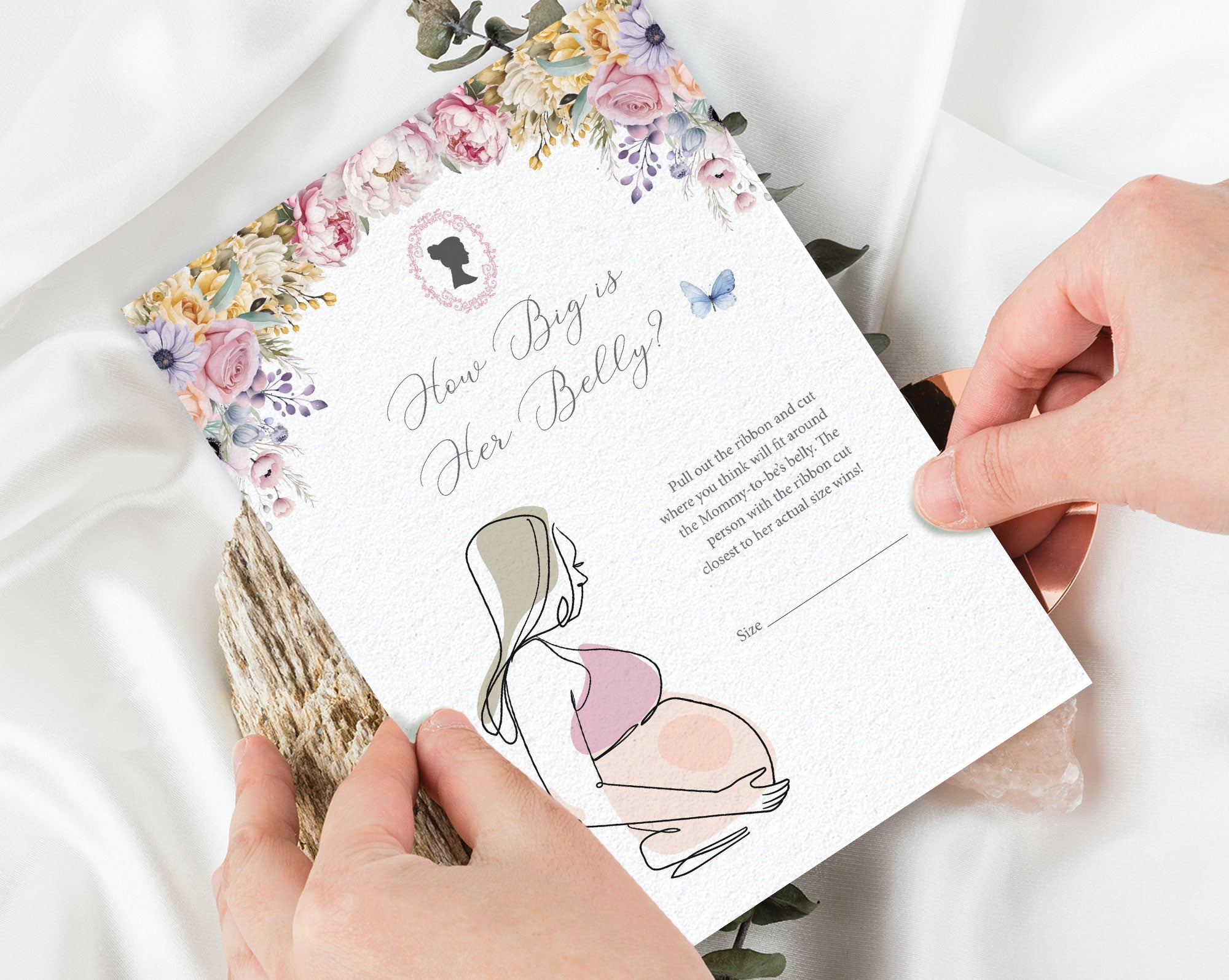 two hands holding a 'how big is her belly' baby shower game card with a white silk cloth in the background