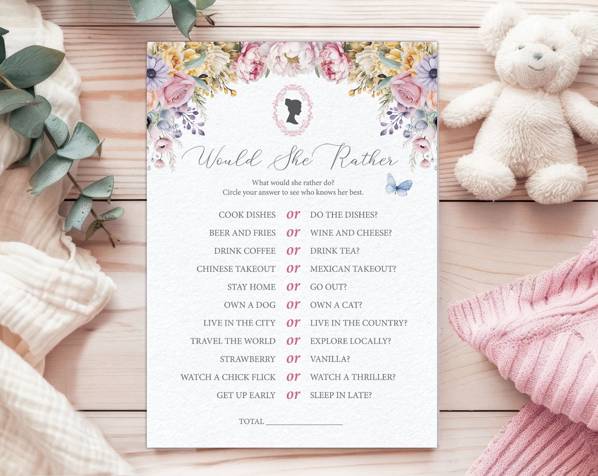 A baby shower game card titled 'would she rather' laying on a wooden background, next to a white teddy bear, a pink and a white blanket and some eucalyptus leaves