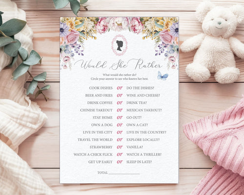 A baby shower game card titled 'would she rather' laying on a wooden background, next to a white teddy bear, a pink and a white blanket and some eucalyptus leaves