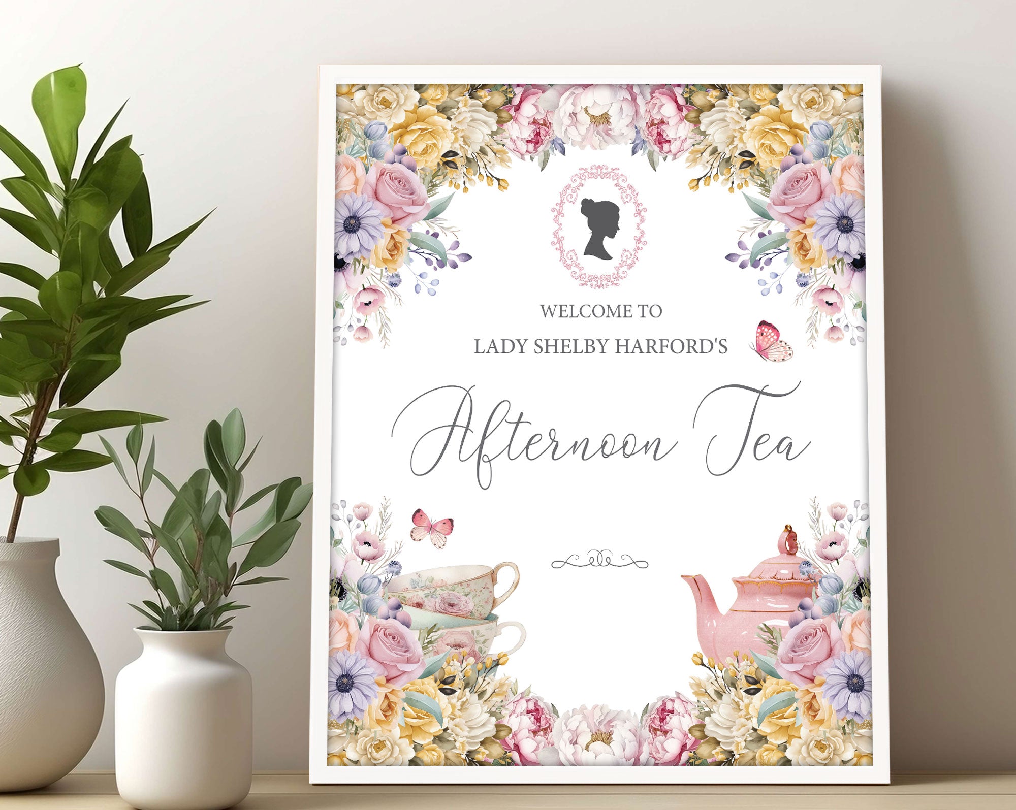 Decorative pastel sign for an afternoon tea event with flowers, cameo and teacups, in a white frame leaning up against a beige wall next to greenery in a vase