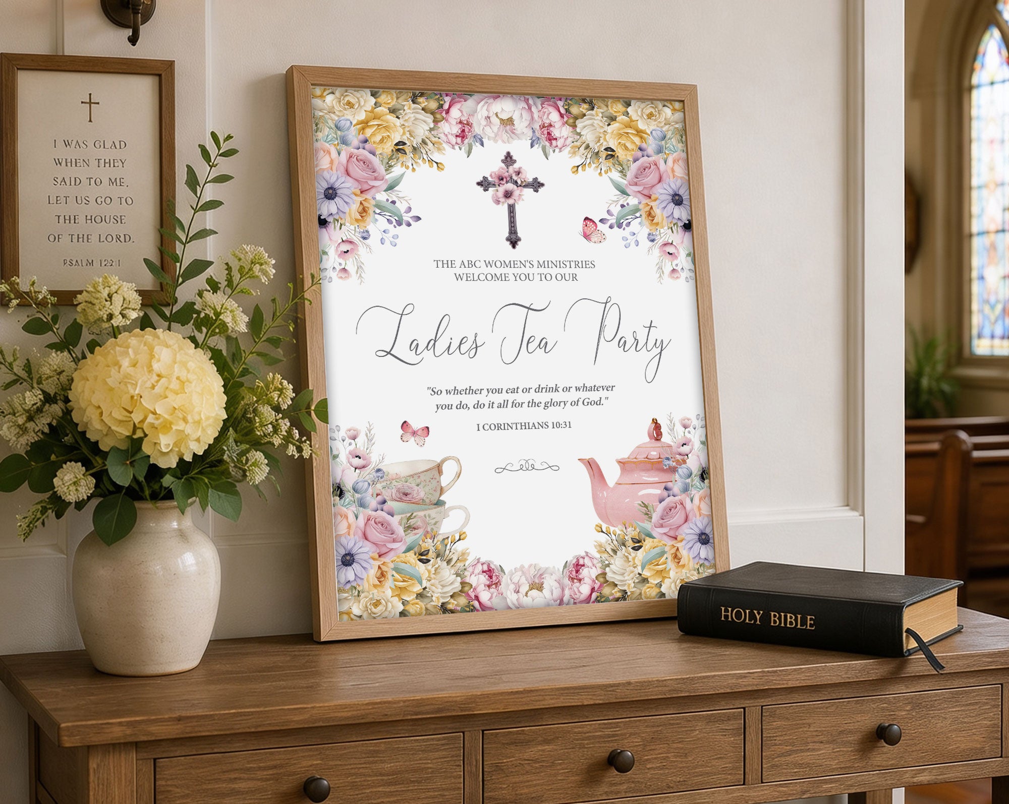 Women's ministries ladies tea party pastel flower welcome sign in a wooden frame on a console table leaning up against a beige wall, with a vase of flowers and a bible next to it