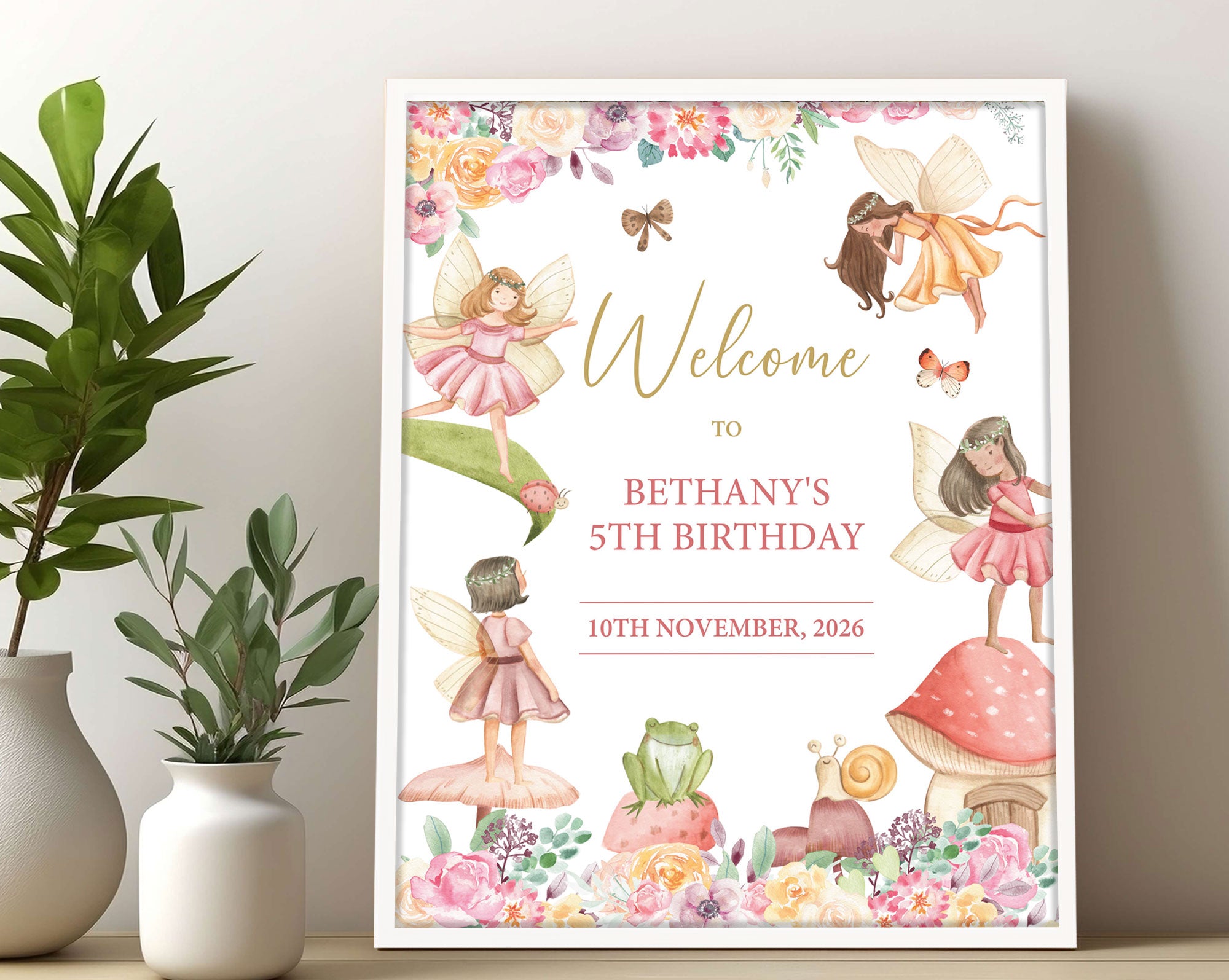 a fairy girls birthday welcome sign with toadstool, frog and girl fairies, in a white frame leaning against a beige wall next to greenery in vases