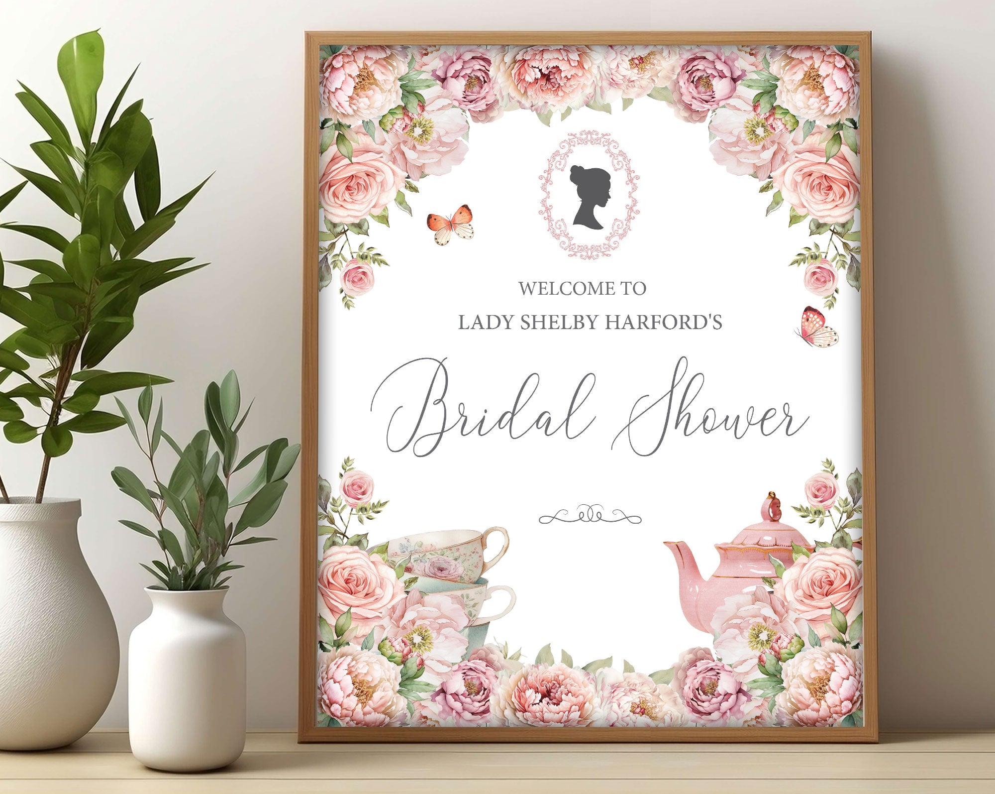 pretty Bridal shower sign with blush pink flowers and regency cameo in a wooden frame, leaning up against a beige wall next to some greenery in vases