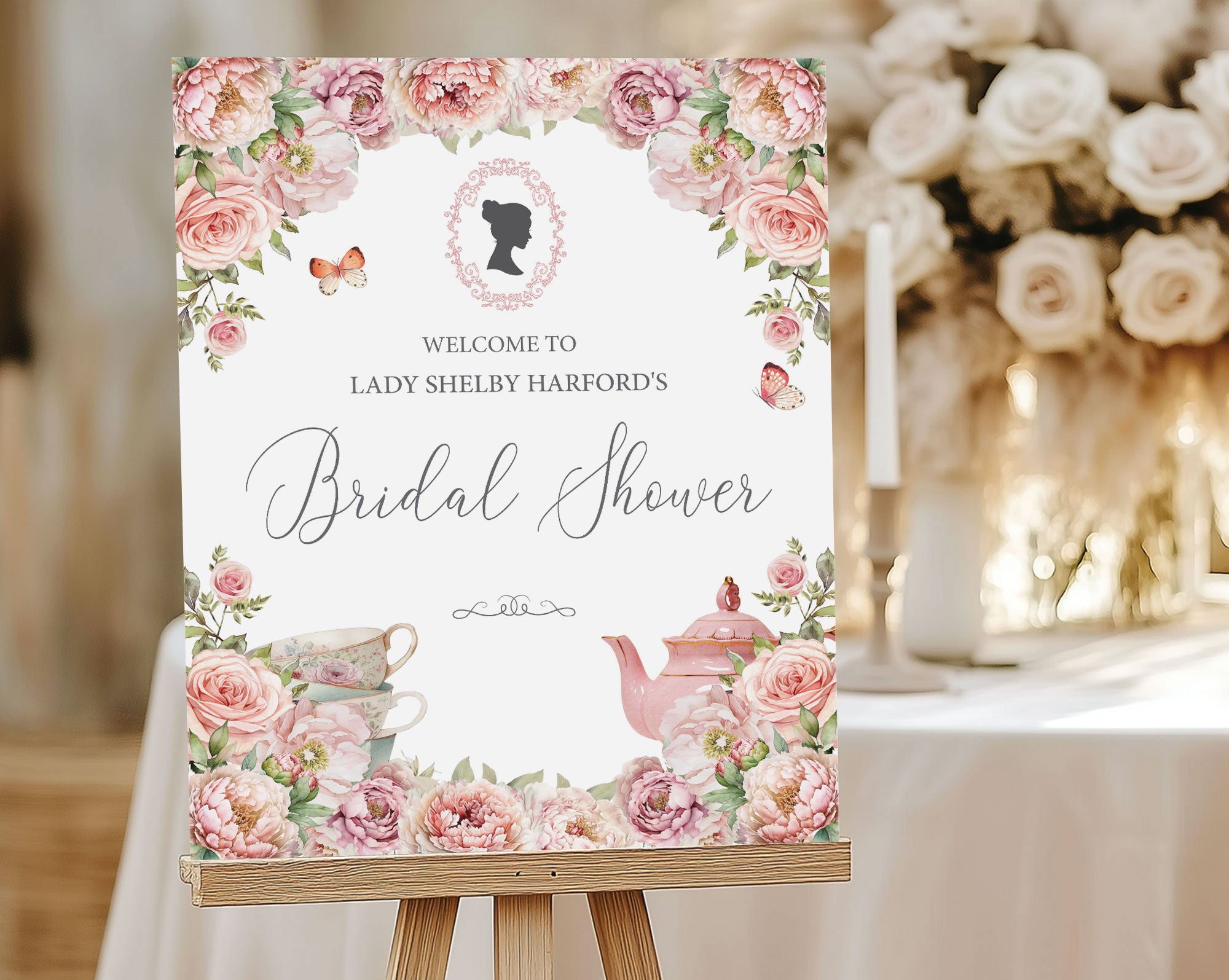 a blush pink flower bridal shower welcome sign with regency cameo, butterflies and tea set graphics, on a wooden easel, in front of a table with candles and cream flowers
