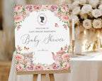 a blush pink flower baby shower welcome sign with regency cameo, butterflies and tea set graphics, on a wooden easel, in front of a table with candles and cream flowers