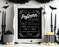 A Halloween-themed sign reading 'The Haunted Popcorn Stand' in an ornate white frame, styled on a tabletop with black candles, popcorn buckets, spiders, bats, and popcorn against a white wall.