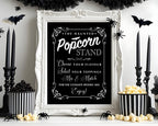 A Halloween-themed sign reading 'The Haunted Popcorn Stand' in an ornate white frame, styled on a tabletop with black candles, popcorn buckets, spiders, bats, and popcorn against a white wall.