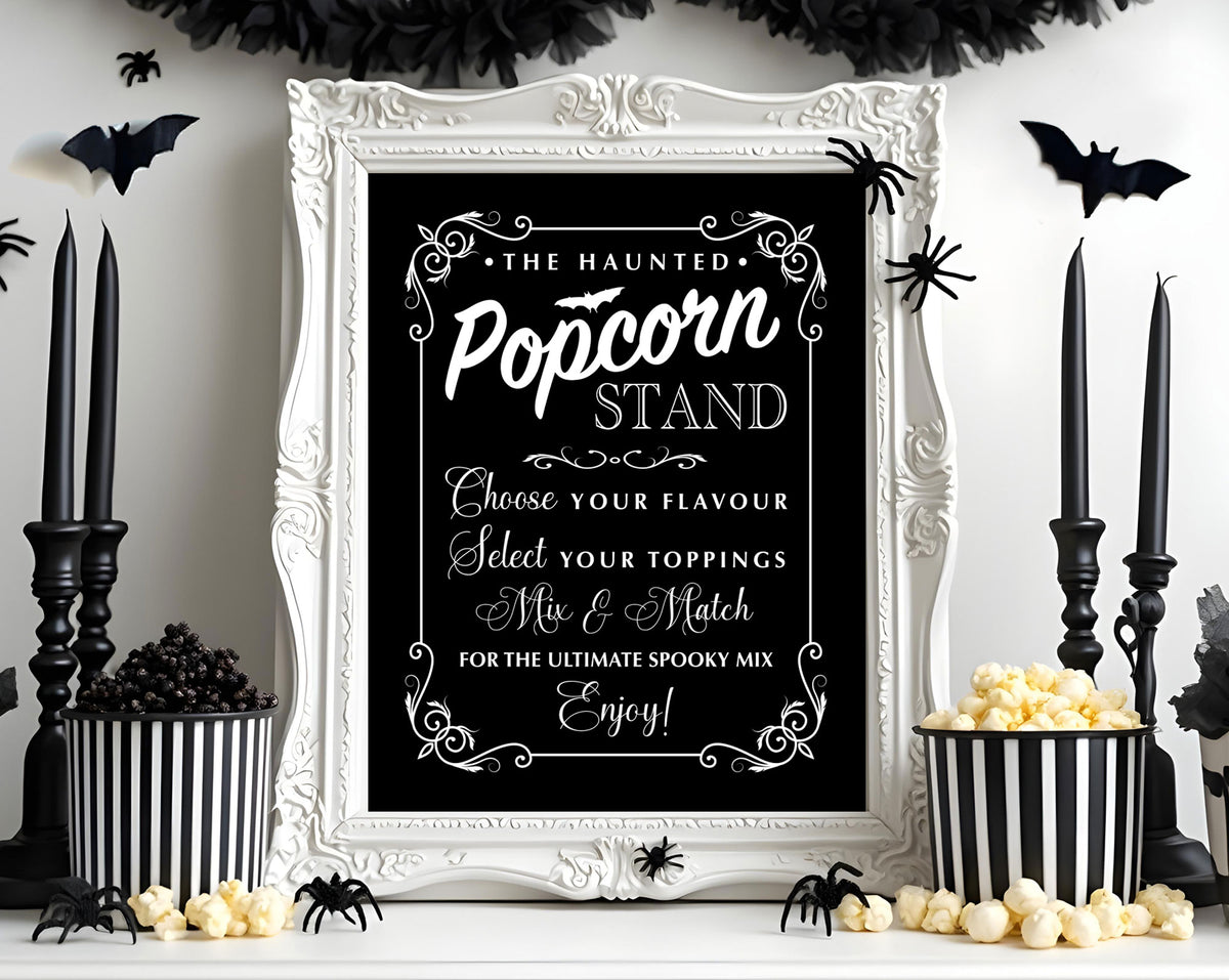 A Halloween-themed sign reading 'The Haunted Popcorn Stand' in an ornate white frame, styled on a tabletop with black candles, popcorn buckets, spiders, bats, and popcorn against a white wall.