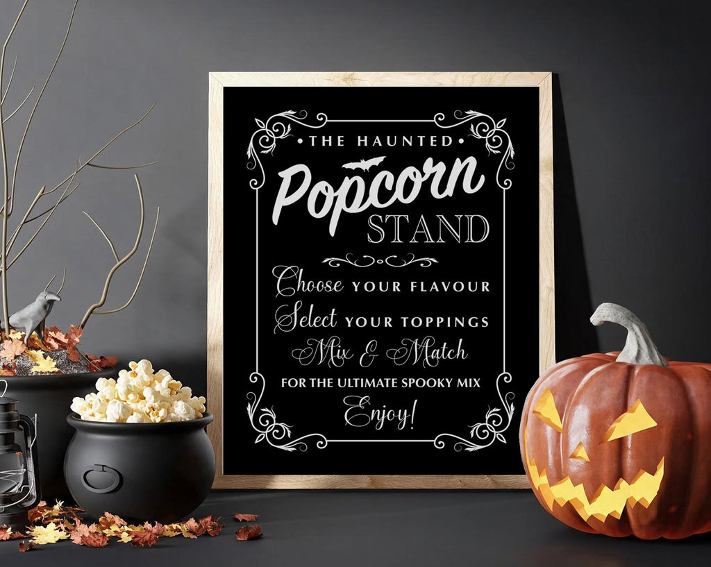 A framed Halloween-themed sign reading 'The Haunted Popcorn Stand' with white decorative text on a black background, styled on a dark tabletop with a pumpkin, popcorn pot, leaves, and branches around it.