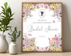 pretty Bridal shower sign with pastel flowers and regency cameo in a wooden frame, leaning up against a beige wall next to some greenery in vases