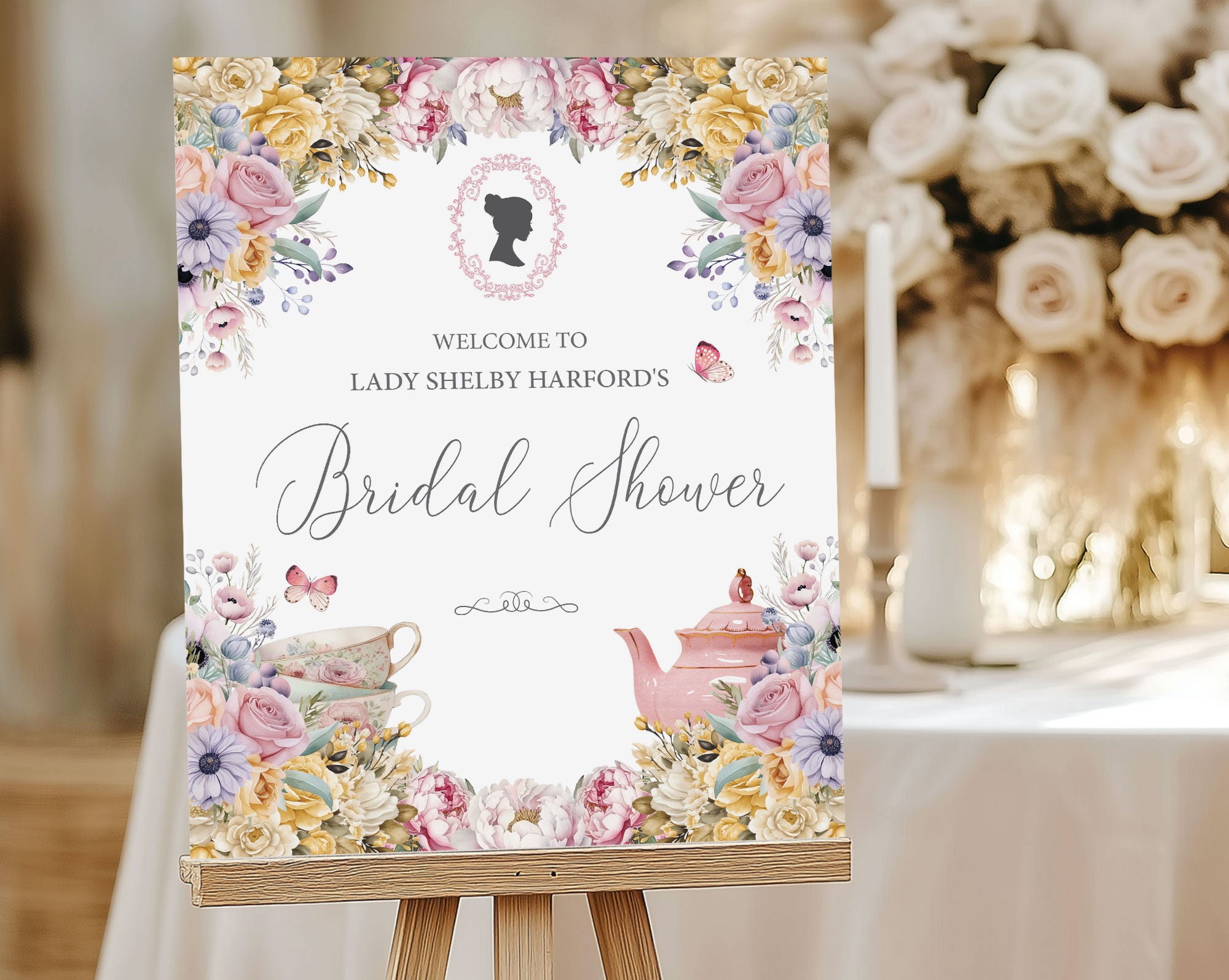 a pastel flower bridal shower welcome sign with regency cameo, butterflies and tea set graphics, on a wooden easel, in front of a table with candles and cream flowers
