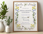 pretty Bridal shower lunch sign with wildflowers, bee and dragonfly in a wooden frame, leaning up against a beige wall next to some greenery in vases