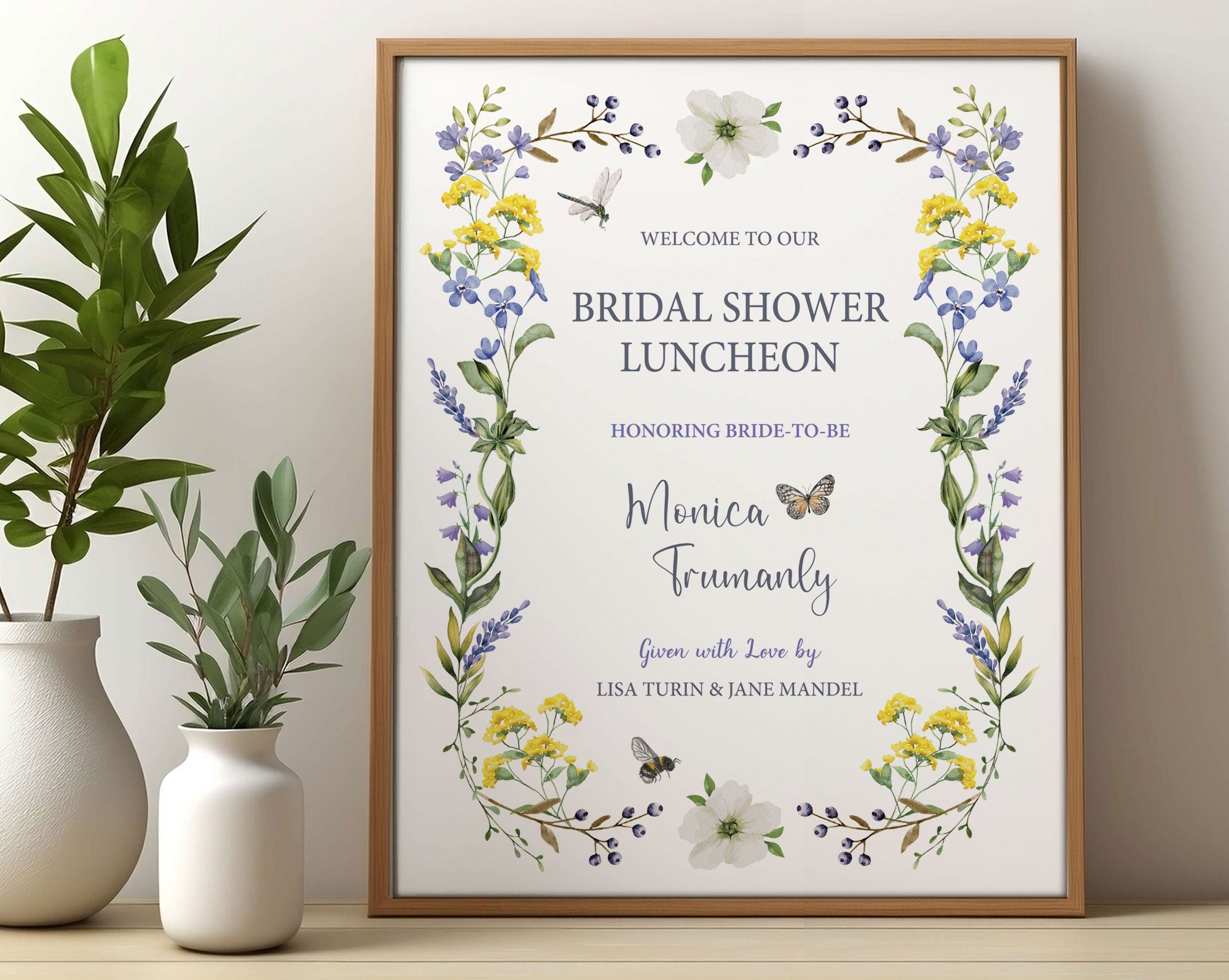 pretty Bridal shower lunch sign with wildflowers, bee and dragonfly in a wooden frame, leaning up against a beige wall next to some greenery in vases