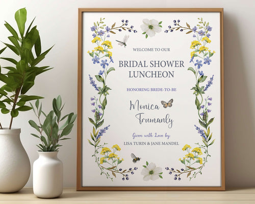 pretty Bridal shower lunch sign with wildflowers, bee and dragonfly in a wooden frame, leaning up against a beige wall next to some greenery in vases