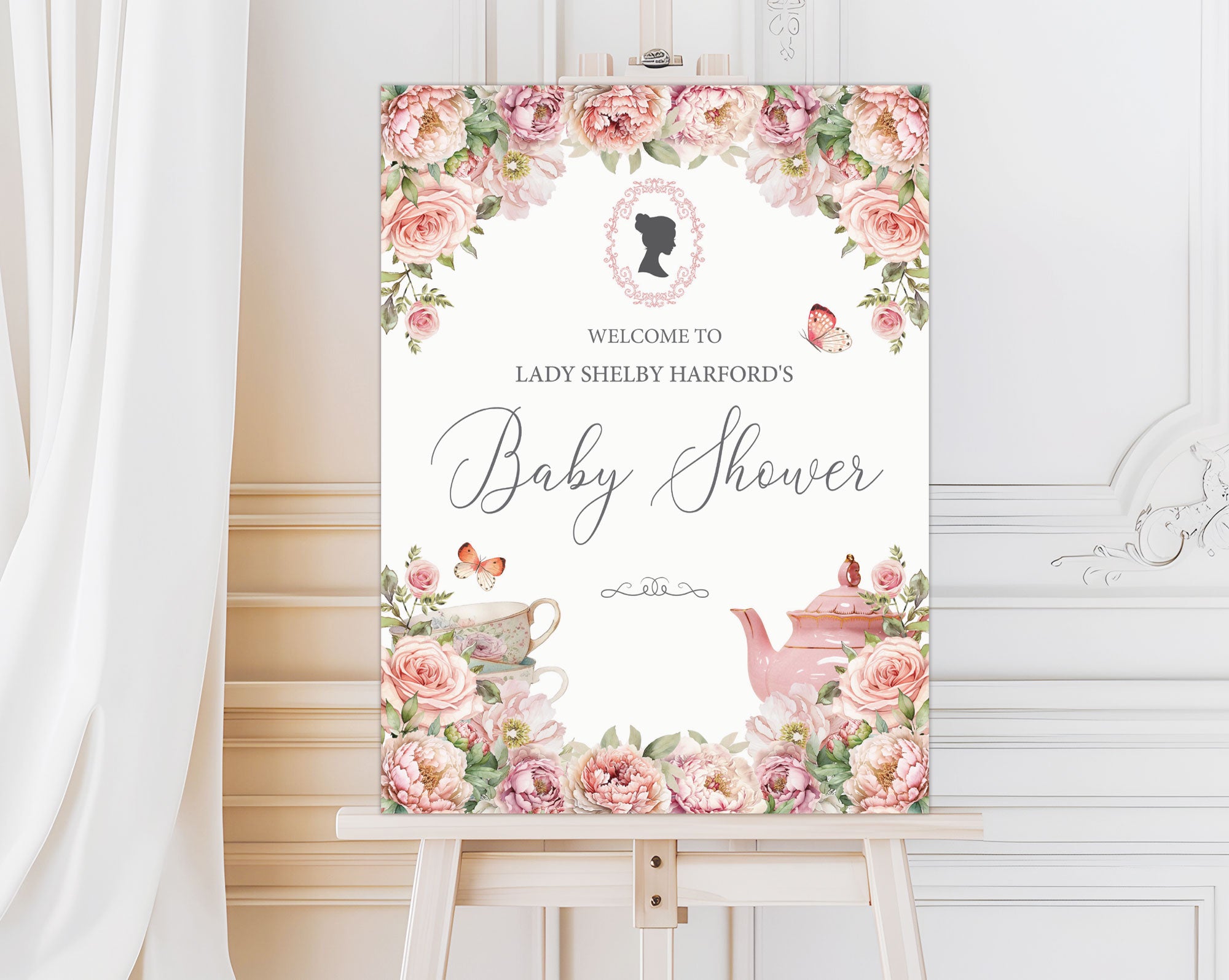 elegant blush pink entrance sign for a baby shower with teapot and cameo on a wooden easel in front of a white panelled wall and curtain