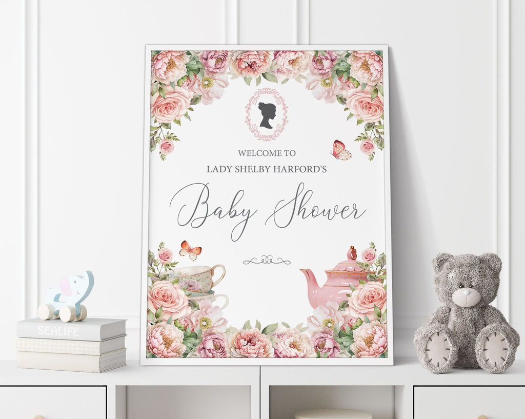 18 x 24 baby shower welcome sign with regency style cameo and blush pink flowers, tea pot and tea cup graphics, in a white frame, leaning up against a white panelled wall next to a grey teddy bear and some books