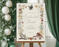 an enchanted 24 x 36 welcome sign with a recycled finish, raven, woodland animals and flora & fauna, on a wooden easel in front of a green curtain next to some greenery with white balloons