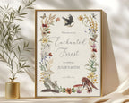 enchanted 18 x 24 sign in a gold frame with raven, forest animals and flora and fauna, leaning up against a beige wall next to some greenery in a gold vase