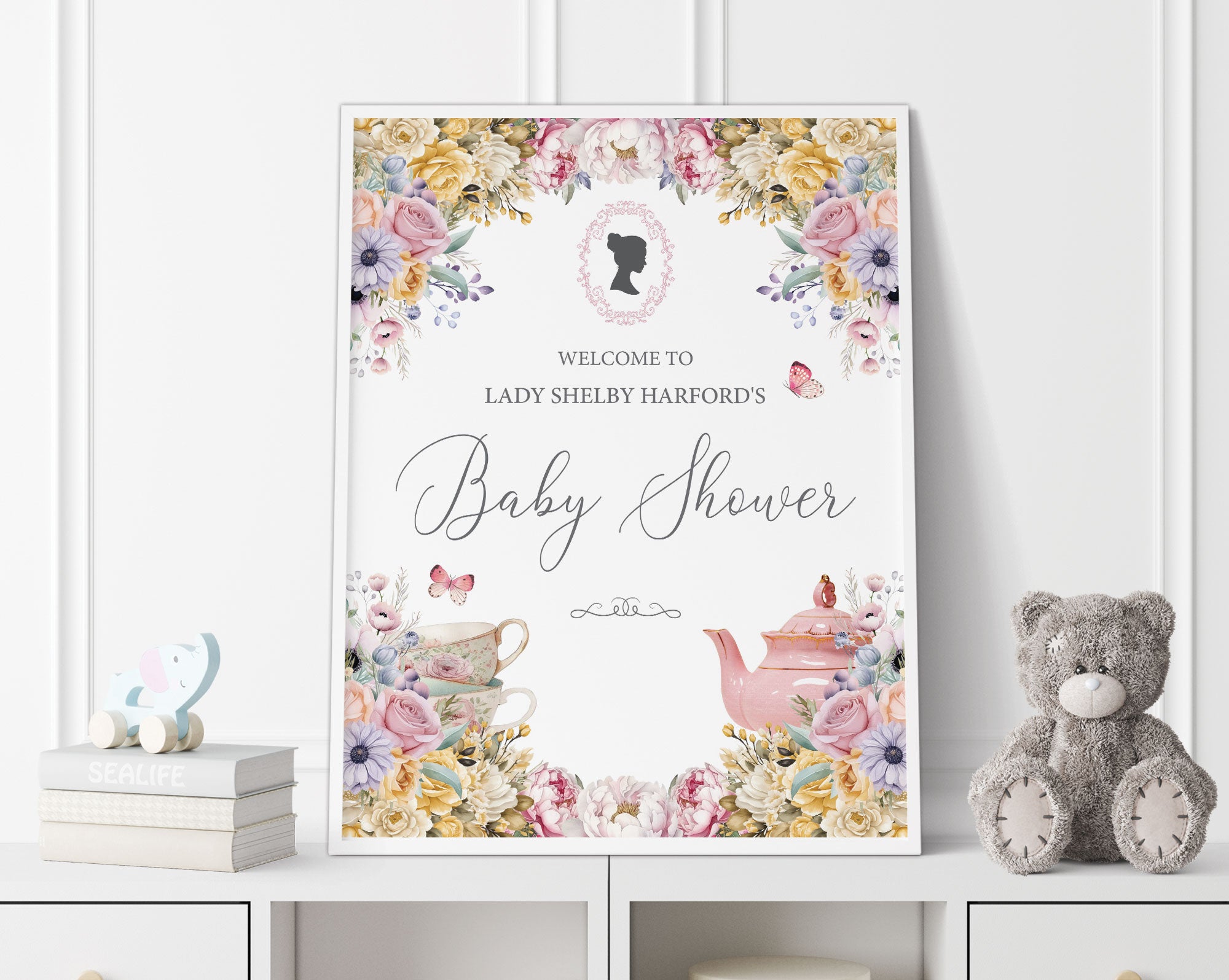 18 x 24 baby shower welcome sign with regency style cameo and pastel flowers, tea pot and tea cup graphics, in a white frame, leaning up against a white panelled wall next to a grey teddy bear and some books