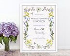 pretty blue purple and yellow flower bridal shower luncheon welcome sign with dragonfly, bee and butterfly graphics, in a white frame, leaning up against a white wall, next to a vase with purple flowers