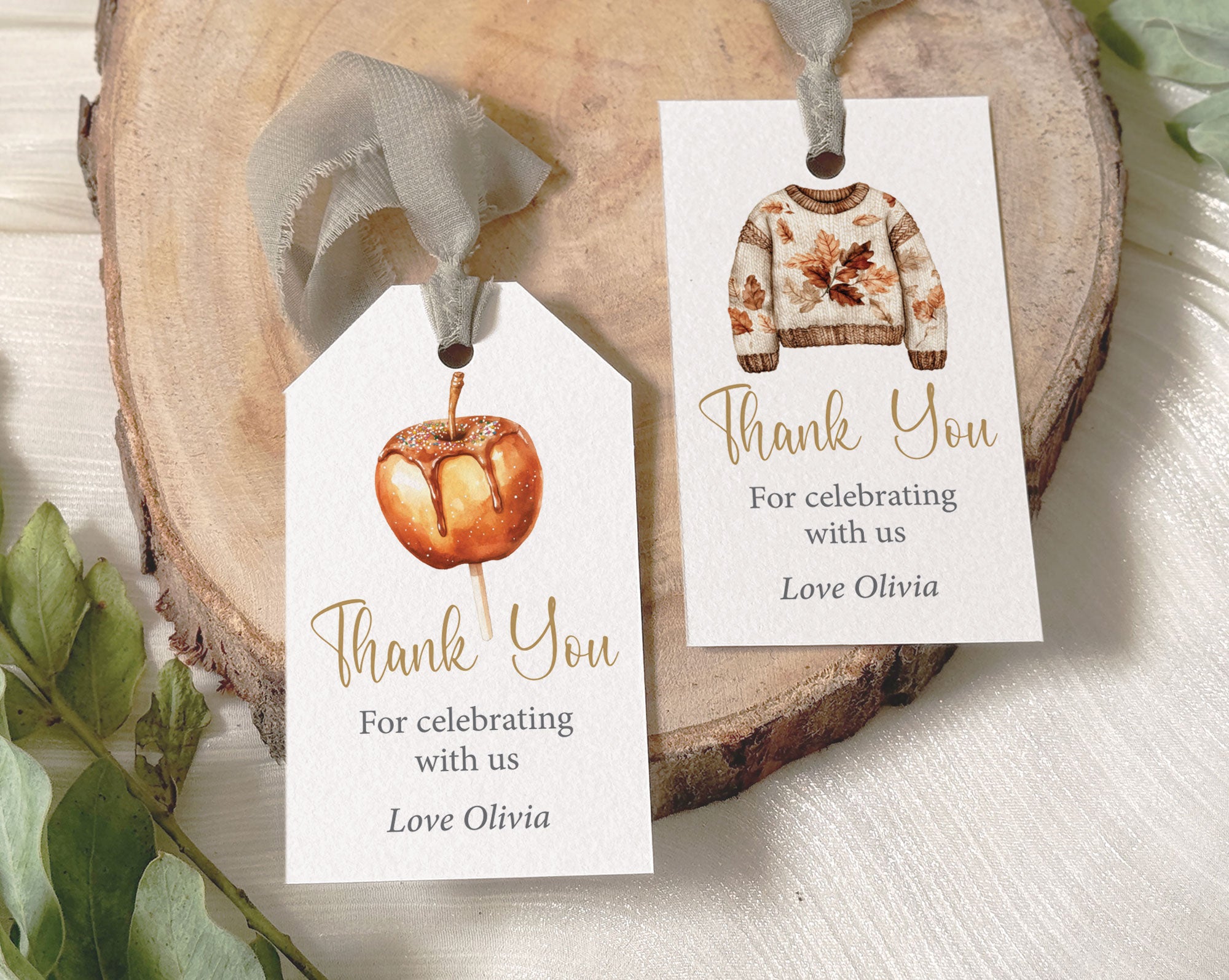 Two personalized thank you favor tags with autumn-themed illustrations, displayed on a rustic wood slice with green leaves. One tag features a watercolor pumpkin, and the other features a watercolor knit sweater.