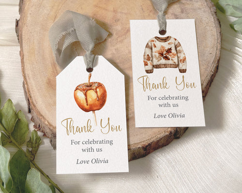 Two personalized thank you favor tags with autumn-themed illustrations, displayed on a rustic wood slice with green leaves. One tag features a watercolor pumpkin, and the other features a watercolor knit sweater.