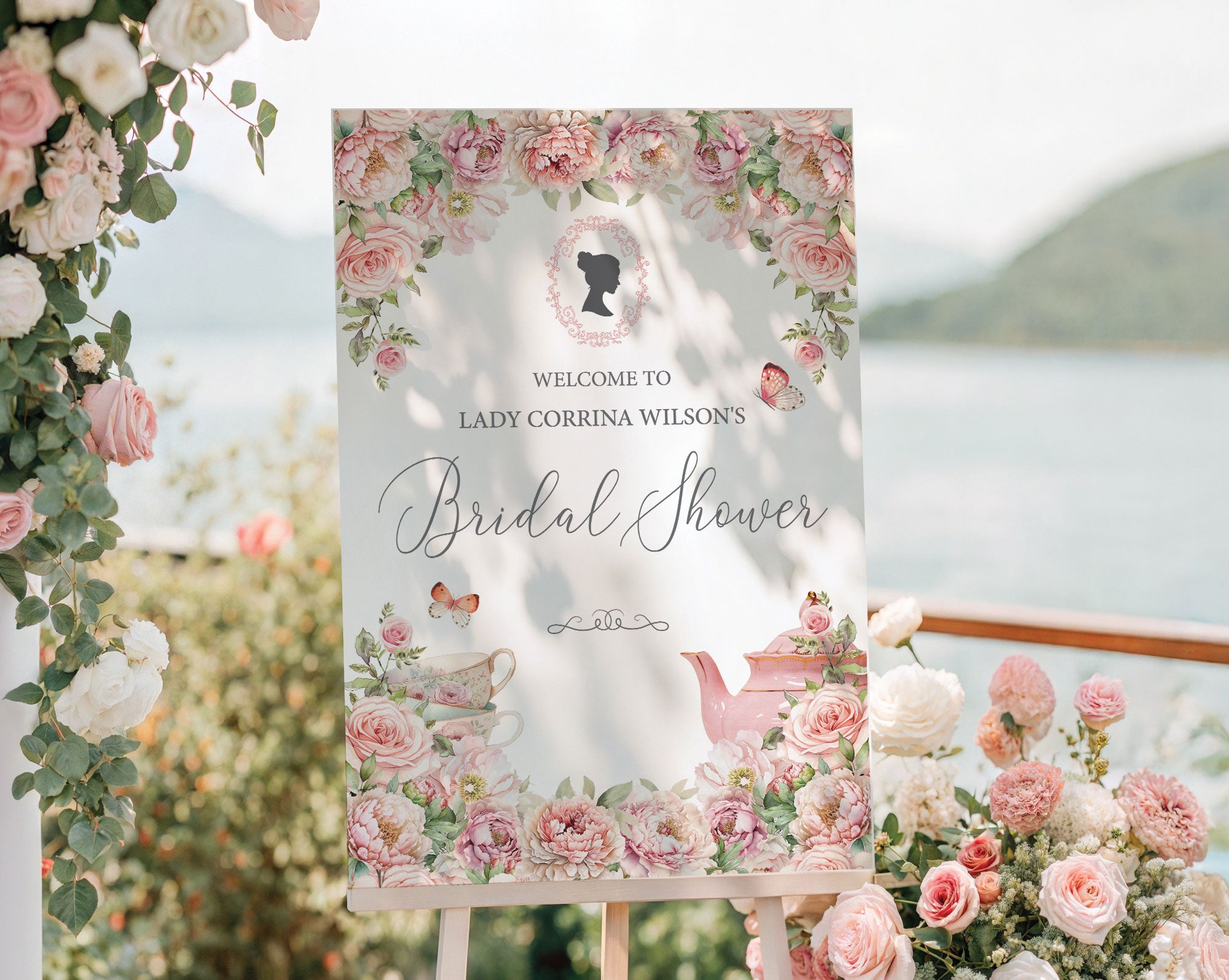 pretty pink flower welcome sign for a bridal shower, outside on a wooden easel in front of a lake backdrop next to trailing vines with pink and white flowers