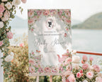 pretty pink flower welcome sign for a baby shower, outside on a wooden easel in front of a lake backdrop next to trailing vines with pink and white flowers