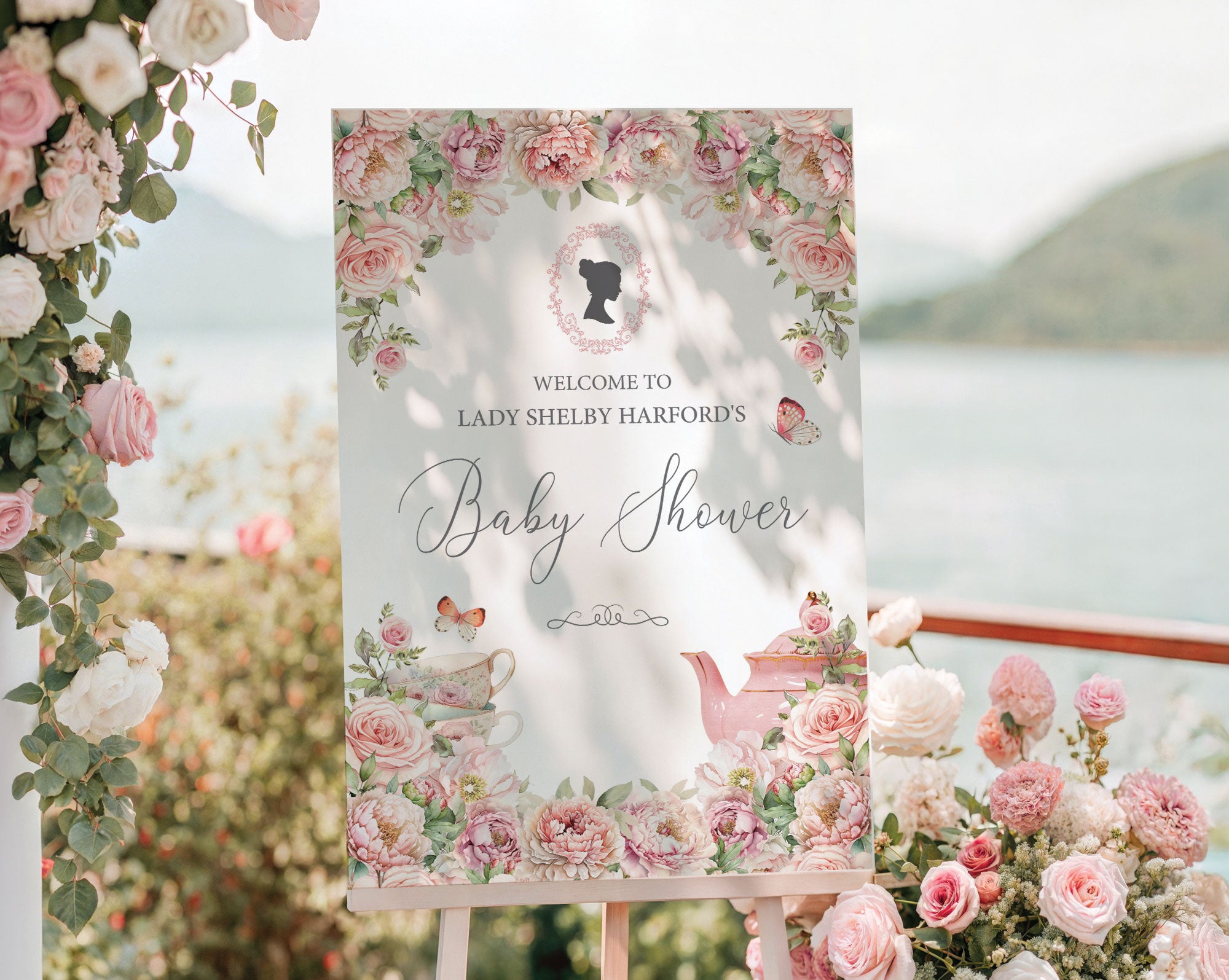 pretty pink flower welcome sign for a baby shower, outside on a wooden easel in front of a lake backdrop next to trailing vines with pink and white flowers
