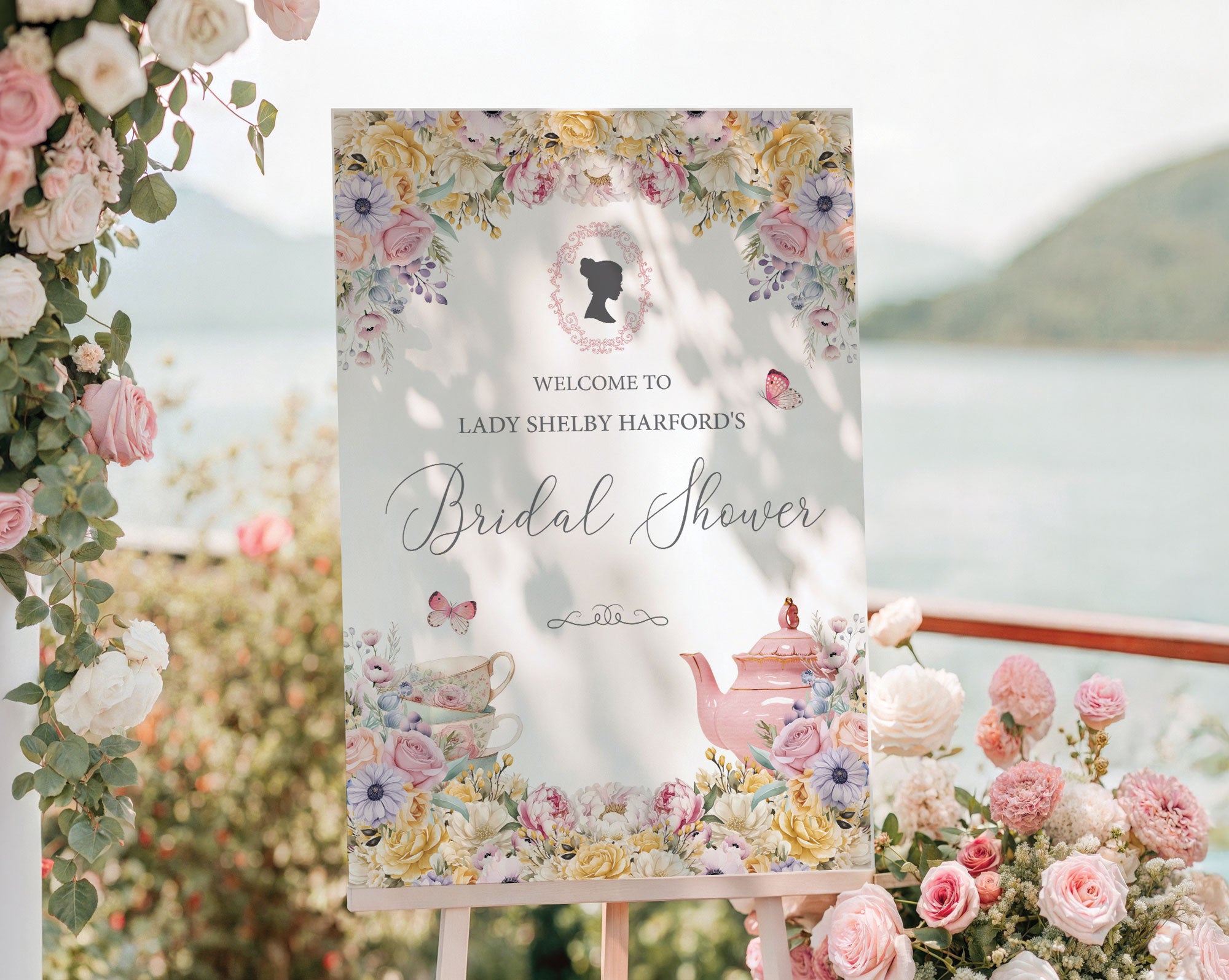 pretty pastel pink and yellow flower welcome sign for a bridal shower, outside on a wooden easel in front of a lake backdrop next to trailing vines with pink and white flowers