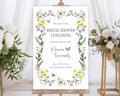 yellow purple wildflower bridal shower welcome sign with bees, dragonfly and butterfly on a wooden easel in front of a white curtain and some flowers on stands