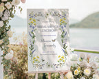 pretty purple and yellow wildflower welcome sign for a bridal shower, outside on a wooden easel in front of a lake backdrop next to trailing vines with yellow and white flowers