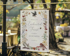 a enchanted forest welcome sign in a gold frame hanging outside in front of a forest setting with tables and chairs in the background