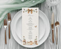 A pretty vertical menu card with a rustic autumn theme, styled on a white plate with rose gold cutlery, a sage green napkin, and a glass on a gray wood table.