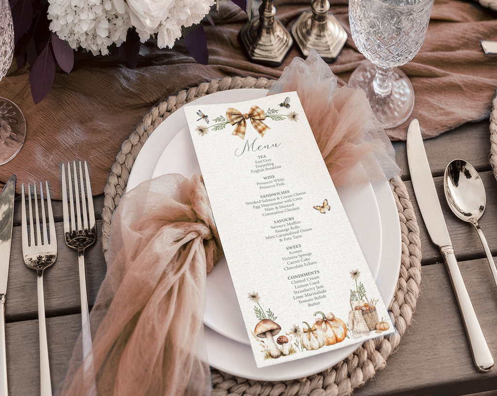 A styled fall / Autumn table setting featuring a vertical menu card with watercolor illustrations of pumpkins, a bow, and butterflies, placed on a white plate with a brown tulle napkin and woven placemat.