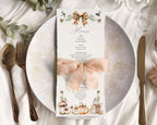 A vertical dinner menu card with autumn-themed illustrations and text, tied with a pale apricot ribbon and bow, displayed on a white plate with silverware and a glass in a styled tabletop setting.
