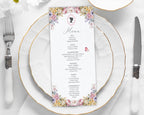 Menu card with floral and cameo design on a white plate with gold rim, silverware, and white flowers.
