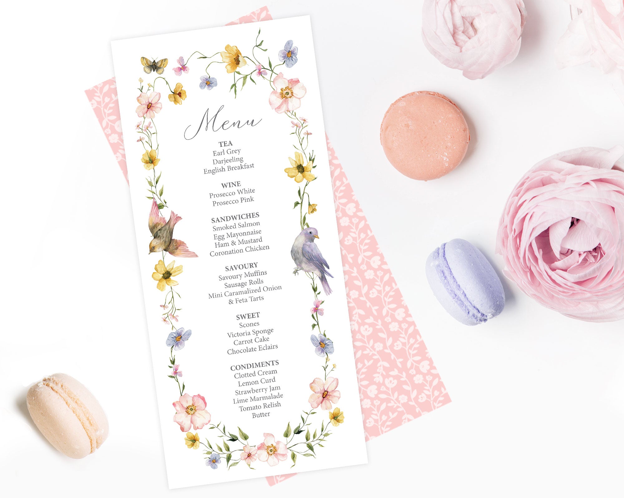 Floral menu card with birds and butterfly on a white surface with macarons and flowers.